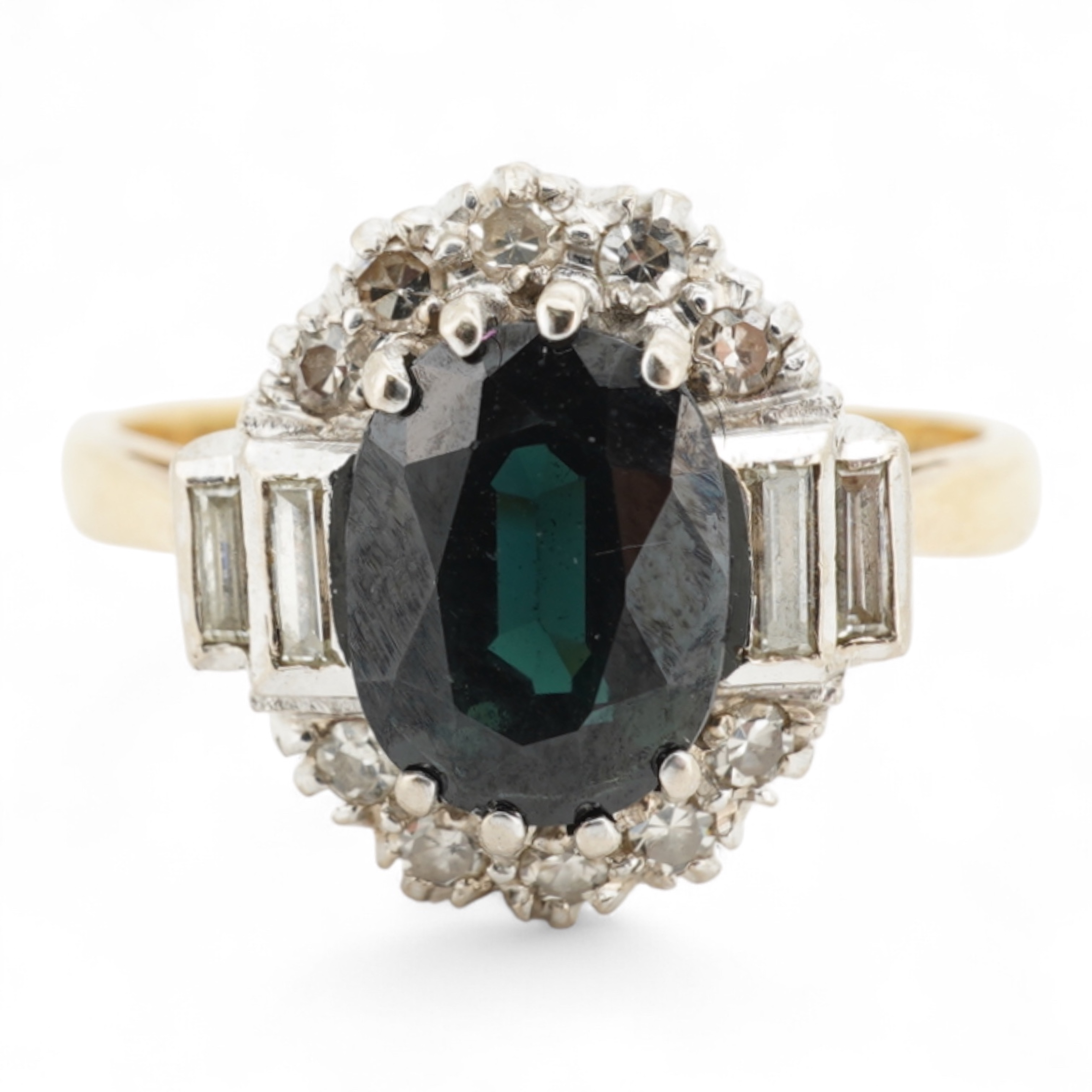 A sapphire and diamond cluster ring, claw-set with an oval sapphire measuring approximately 8.2 x 6.1 x 4.3mm, within a border of brilliant-cut and baguette diamonds totalling approximately 0.20 carats, mounted in 18ct g